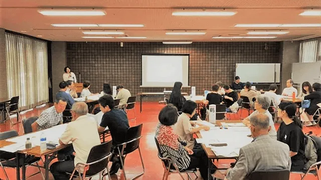 写真：各テーブルに分かれて座った二十人程度の方達。壇上でマイクを持った方が話している。